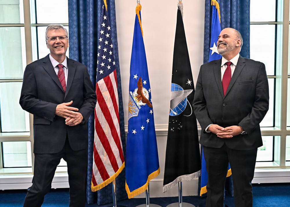 Two men in dark business suits with red ties are posing together in what appears to be an official government setting. They are smiling and standing in front of multiple flags, including the United States flag, the U.S. Air Force flag, and the U.S. Space Force flag. The setting suggests a formal diplomatic or military meeting.