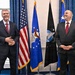 Two men in dark business suits with red ties are posing together in what appears to be an official government setting. They are smiling and standing in front of multiple flags, including the United States flag, the U.S. Air Force flag, and the U.S. Space Force flag. The setting suggests a formal diplomatic or military meeting.