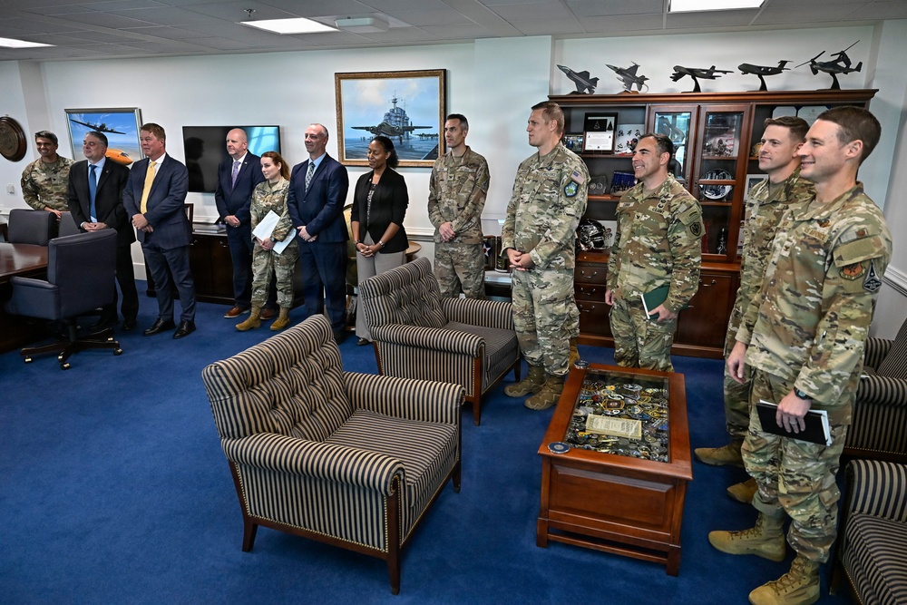 A formal gathering in what appears to be a senior military officer's office, featuring a mix of uniformed military personnel and civilian officials standing together. The setting includes aviation-themed decorations, aircraft models on shelving units, military artwork, and a glass-topped coffee table displaying challenge coins. The group appears to be assembled for an official meeting, briefing, or recognition ceremony.