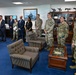 A formal gathering in what appears to be a senior military officer's office, featuring a mix of uniformed military personnel and civilian officials standing together. The setting includes aviation-themed decorations, aircraft models on shelving units, military artwork, and a glass-topped coffee table displaying challenge coins. The group appears to be assembled for an official meeting, briefing, or recognition ceremony.