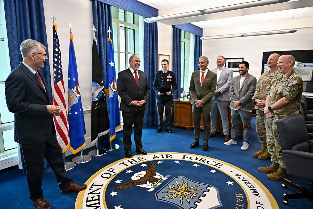 A group of approximately eight individuals are gathered in what appears to be a formal office within the Department of the Air Force, standing on a large official seal rug. The group includes civilians in business attire, one uniformed military officer in dress blues, and two individuals in Army combat uniforms. The atmosphere appears cordial and informal, with several people smiling and laughing.