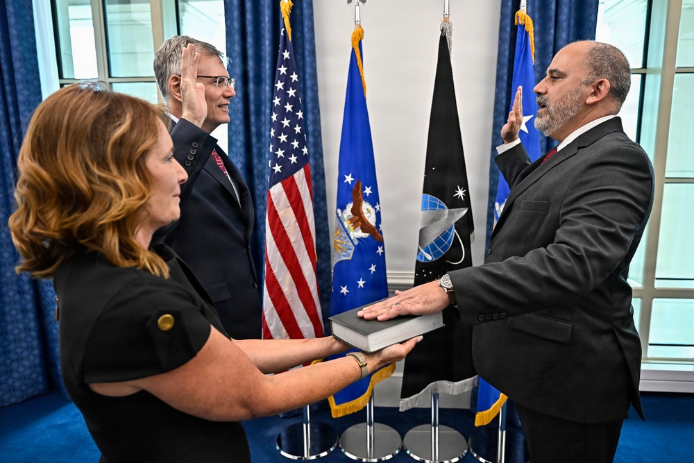 A formal swearing-in ceremony is taking place in what appears to be a government office setting. A man in a dark suit places his hand on a book held by a woman while raising his right hand, being administered an oath by another official. The backdrop includes the American flag and what appear to be Air Force and Space Force flags.