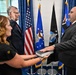 A formal swearing-in ceremony is taking place in what appears to be a government office setting. A man in a dark suit places his hand on a book held by a woman while raising his right hand, being administered an oath by another official. The backdrop includes the American flag and what appear to be Air Force and Space Force flags.