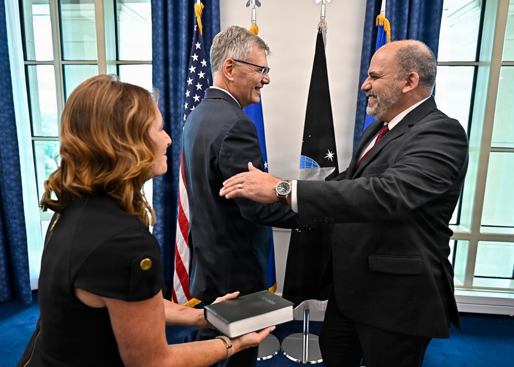 Three individuals are gathered in what appears to be an official government ceremony, likely a swearing-in or oath-taking event. Two men in dark suits are shaking hands and laughing, while a woman in a black dress holds a Bible or official book. The setting includes a U.S. flag, an Air Force flag, and what appears to be a Space Force flag in the background.