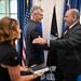 Three individuals are gathered in what appears to be an official government ceremony, likely a swearing-in or oath-taking event. Two men in dark suits are shaking hands and laughing, while a woman in a black dress holds a Bible or official book. The setting includes a U.S. flag, an Air Force flag, and what appears to be a Space Force flag in the background.