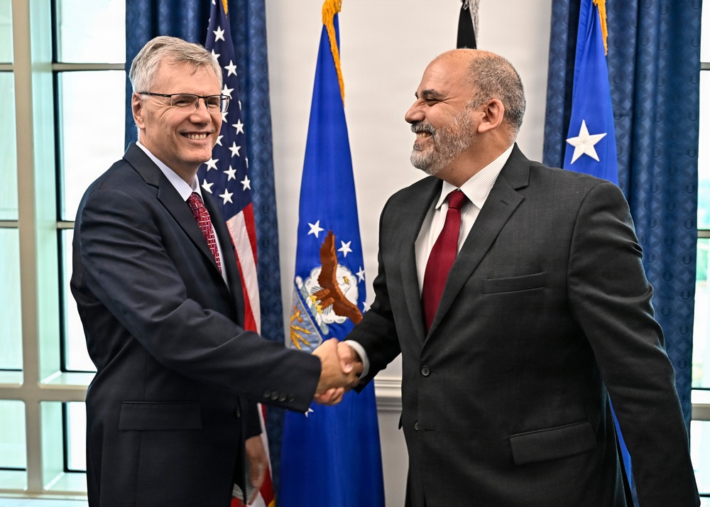 Two men in dark business suits are shaking hands and smiling in what appears to be an official government setting. Both are wearing red ties, suggesting a formal diplomatic or official meeting. The background features the United States flag and what appears to be a U.S. Air Force flag, indicating a military or government venue.