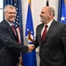 Two men in dark business suits are shaking hands and smiling in what appears to be an official government setting. Both are wearing red ties, suggesting a formal diplomatic or official meeting. The background features the United States flag and what appears to be a U.S. Air Force flag, indicating a military or government venue.