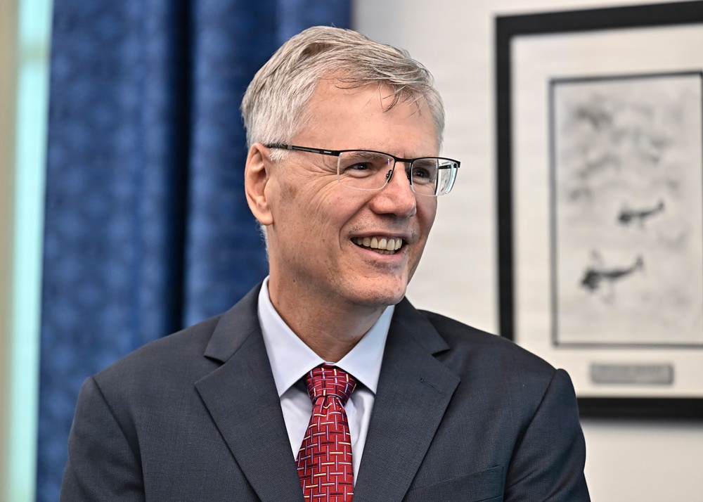 A middle-aged man with gray hair and rectangular-framed glasses is smiling broadly in what appears to be a professional or office setting. He is dressed formally in a gray suit, white dress shirt, and a red patterned tie. A framed black-and-white artwork or print is visible on the wall behind him.
