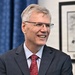 A middle-aged man with gray hair and rectangular-framed glasses is smiling broadly in what appears to be a professional or office setting. He is dressed formally in a gray suit, white dress shirt, and a red patterned tie. A framed black-and-white artwork or print is visible on the wall behind him.