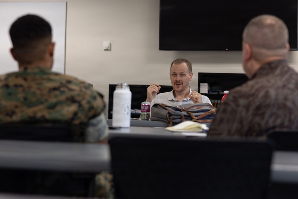 A civilian man in a light-colored polo shirt appears to be speaking or presenting to two individuals seated with their backs to the camera. One of the listeners is wearing U.S. Marine Corps MARPAT camouflage uniform, while the other wears a brown patterned civilian shirt. The setting appears to be a conference or briefing room with a wall-mounted TV screen visible in the background.