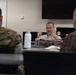 A civilian man in a light-colored polo shirt appears to be speaking or presenting to two individuals seated with their backs to the camera. One of the listeners is wearing U.S. Marine Corps MARPAT camouflage uniform, while the other wears a brown patterned civilian shirt. The setting appears to be a conference or briefing room with a wall-mounted TV screen visible in the background.