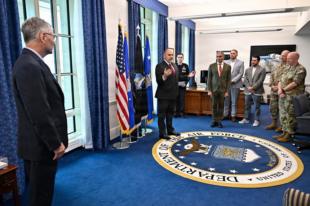 A formal meeting or briefing is taking place in what appears to be a high-level U.S. Department of the Air Force office, identifiable by the large official seal embedded in the blue carpet. A man in a dark suit appears to be speaking or presenting to a group of mixed civilian and military personnel. The setting includes official flags and formal office furnishings consistent with a senior leadership space.