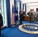 A formal meeting or briefing is taking place in what appears to be a high-level U.S. Department of the Air Force office, identifiable by the large official seal embedded in the blue carpet. A man in a dark suit appears to be speaking or presenting to a group of mixed civilian and military personnel. The setting includes official flags and formal office furnishings consistent with a senior leadership space.