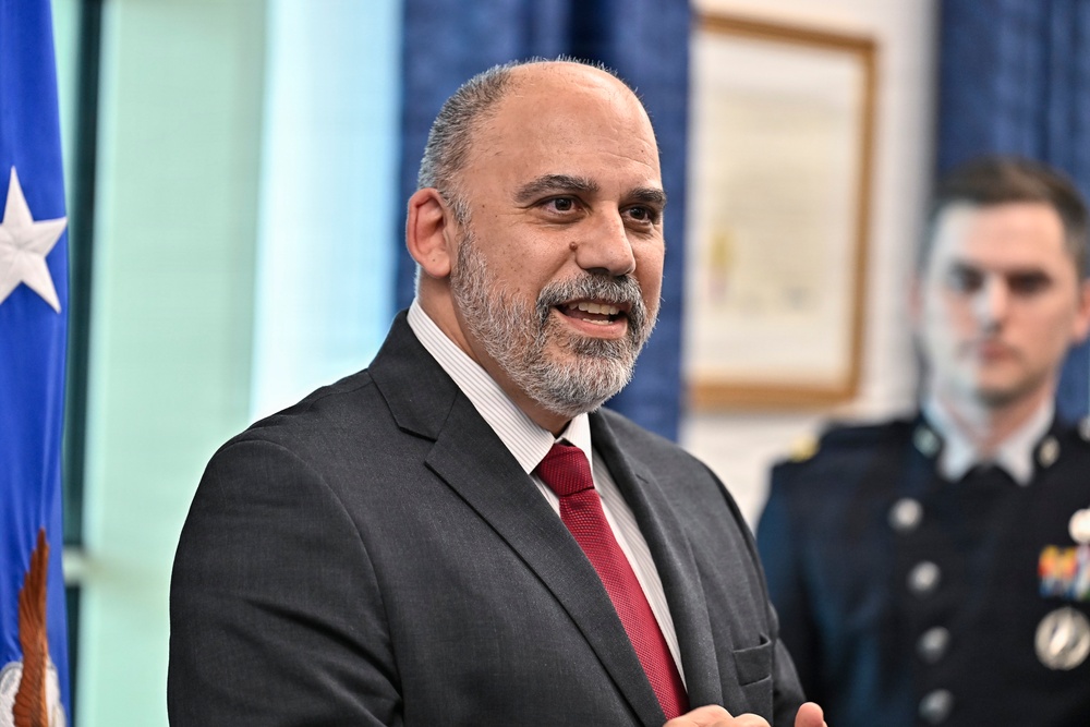 A middle-aged man in a dark gray suit with a red tie is speaking or presenting in what appears to be an official or formal setting. Behind him is a U.S. Air Force flag and a uniformed military personnel member. The setting suggests a formal ceremony or official government/military event.