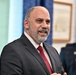 A middle-aged man in a dark gray suit with a red tie is speaking or presenting in what appears to be an official or formal setting. Behind him is a U.S. Air Force flag and a uniformed military personnel member. The setting suggests a formal ceremony or official government/military event.