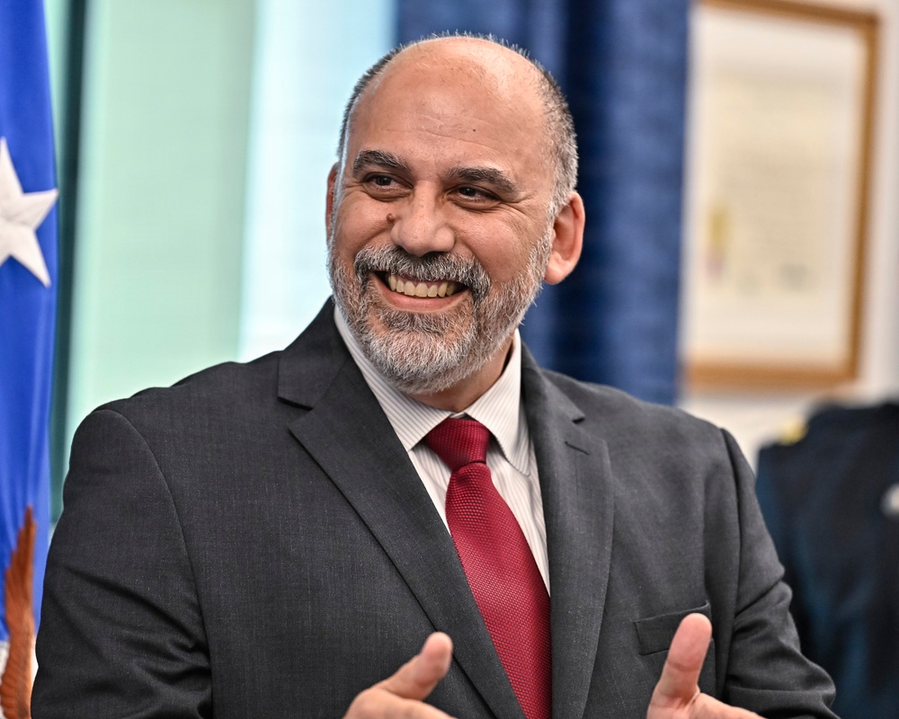 A middle-aged man with a salt-and-pepper beard and receding hairline is smiling broadly and gesturing with his hands, suggesting he is speaking or presenting. He is dressed in a dark gray suit with a red tie and striped dress shirt. The setting appears to be an official or governmental environment, indicated by a flag with stars visible in the background.