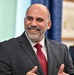 A middle-aged man with a salt-and-pepper beard and receding hairline is smiling broadly and gesturing with his hands, suggesting he is speaking or presenting. He is dressed in a dark gray suit with a red tie and striped dress shirt. The setting appears to be an official or governmental environment, indicated by a flag with stars visible in the background.