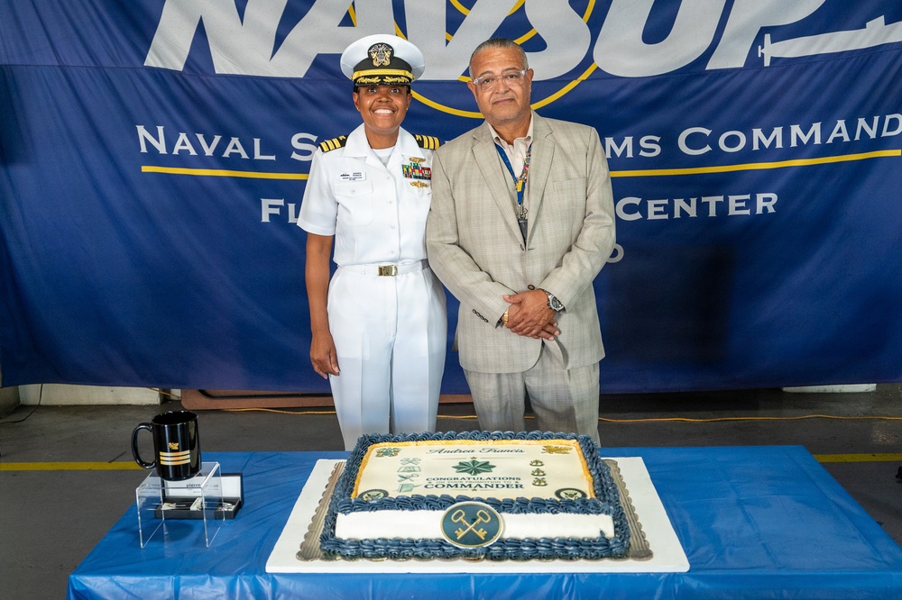 A U.S. Navy officer in white dress uniform stands alongside a civilian man in a light gray suit in front of a NAVSUP (Naval Supply Systems Command) banner. A congratulatory cake on the table reads 'Andrea Francis - Congratulations Commander,' indicating a promotion ceremony. The setting appears to be an official military recognition event at a Fleet Logistics Center.