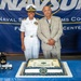 A U.S. Navy officer in white dress uniform stands alongside a civilian man in a light gray suit in front of a NAVSUP (Naval Supply Systems Command) banner. A congratulatory cake on the table reads 'Andrea Francis - Congratulations Commander,' indicating a promotion ceremony. The setting appears to be an official military recognition event at a Fleet Logistics Center.