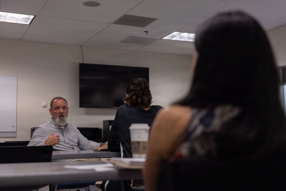 A meeting or classroom discussion is taking place in a conference or seminar room. An older man with a white beard, seated at a table, appears to be speaking or presenting to others in the room. Two individuals with dark hair are visible from behind in the foreground, facing the speaker.