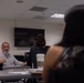 A meeting or classroom discussion is taking place in a conference or seminar room. An older man with a white beard, seated at a table, appears to be speaking or presenting to others in the room. Two individuals with dark hair are visible from behind in the foreground, facing the speaker.