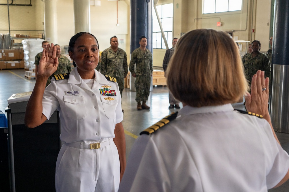 A U.S. Navy officer in white dress uniform is taking an oath, raising her right hand before a senior officer also in white dress uniform. The ceremony appears to be a promotion or re-enlistment ceremony taking place inside a military warehouse or logistics facility. Several sailors in camouflage utility uniforms stand in the background as witnesses.