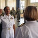 A U.S. Navy officer in white dress uniform is taking an oath, raising her right hand before a senior officer also in white dress uniform. The ceremony appears to be a promotion or re-enlistment ceremony taking place inside a military warehouse or logistics facility. Several sailors in camouflage utility uniforms stand in the background as witnesses.