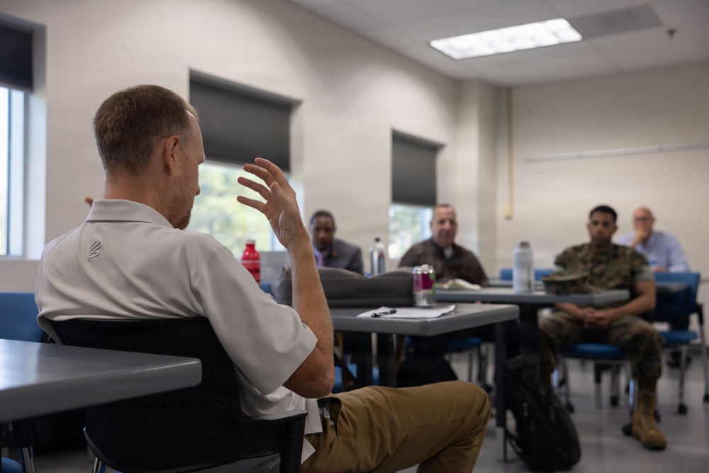 A small group meeting or briefing is taking place in a classroom or conference room setting. A man in a white polo shirt is seated with his back to the camera, gesturing with his raised hand as he speaks to a group of approximately four individuals seated at tables in front of him. At least two of the attendees appear to be wearing military uniforms.