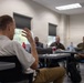 A small group meeting or briefing is taking place in a classroom or conference room setting. A man in a white polo shirt is seated with his back to the camera, gesturing with his raised hand as he speaks to a group of approximately four individuals seated at tables in front of him. At least two of the attendees appear to be wearing military uniforms.