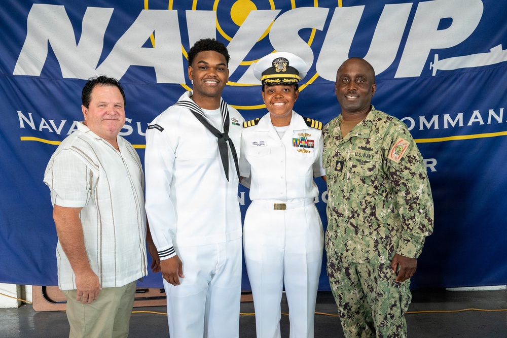 Four individuals pose for a group photo in front of a NAVSUP (Naval Supply Systems Command) banner. Three of the four are wearing U.S. Navy uniforms of varying ranks and types, while one civilian is also present. The setting appears to be an official military event or ceremony.