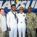 Four individuals pose for a group photo in front of a NAVSUP (Naval Supply Systems Command) banner. Three of the four are wearing U.S. Navy uniforms of varying ranks and types, while one civilian is also present. The setting appears to be an official military event or ceremony.