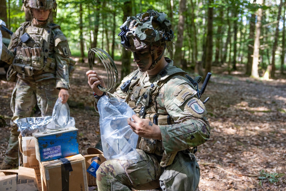 Two military personnel in camouflage uniforms and tactical gear are working in a wooded area with what appears to be demolition or EOD training equipment. The foreground soldier, wearing face paint and a camouflage-covered helmet with night vision mount, is handling coiled wire and a clear plastic bag containing materials. Cardboard boxes labeled 'DEMOLITION KIT REPLICA' are visible nearby, indicating this is a training exercise.