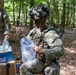 Two military personnel in camouflage uniforms and tactical gear are working in a wooded area with what appears to be demolition or EOD training equipment. The foreground soldier, wearing face paint and a camouflage-covered helmet with night vision mount, is handling coiled wire and a clear plastic bag containing materials. Cardboard boxes labeled 'DEMOLITION KIT REPLICA' are visible nearby, indicating this is a training exercise.