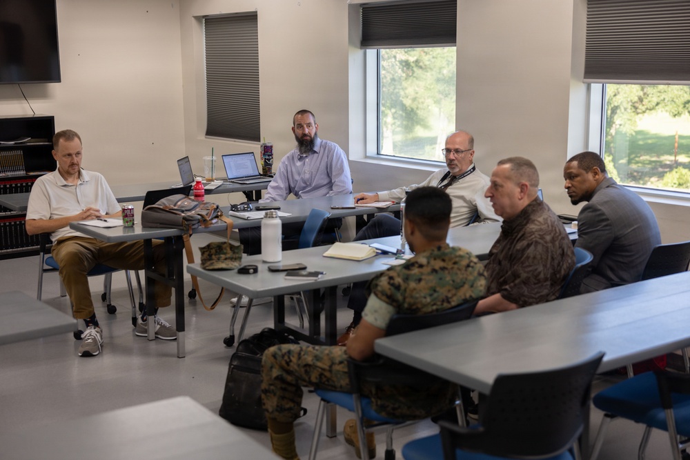 A group of approximately six individuals are seated around tables in a modern classroom or conference room setting. The group includes at least two individuals in military camouflage uniforms (appearing to be U.S. Marine Corps MARPAT pattern) and several civilians. They appear to be engaged in a meeting, briefing, or discussion session.