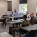 A group of approximately six individuals are seated around tables in a modern classroom or conference room setting. The group includes at least two individuals in military camouflage uniforms (appearing to be U.S. Marine Corps MARPAT pattern) and several civilians. They appear to be engaged in a meeting, briefing, or discussion session.