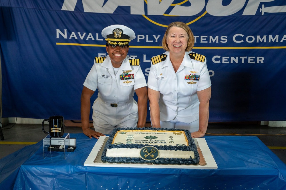 Two U.S. Navy officers in white dress uniforms pose together at a ceremony in front of a NAVSUP (Naval Supply Systems Command) Fleet Logistics Center San Diego banner. A congratulatory cake inscribed with 'Andrea Frances - Congratulations Commander' is displayed on a blue-draped table in front of them. The event appears to be a promotion or change of command ceremony.