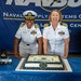 Two U.S. Navy officers in white dress uniforms pose together at a ceremony in front of a NAVSUP (Naval Supply Systems Command) Fleet Logistics Center San Diego banner. A congratulatory cake inscribed with 'Andrea Frances - Congratulations Commander' is displayed on a blue-draped table in front of them. The event appears to be a promotion or change of command ceremony.