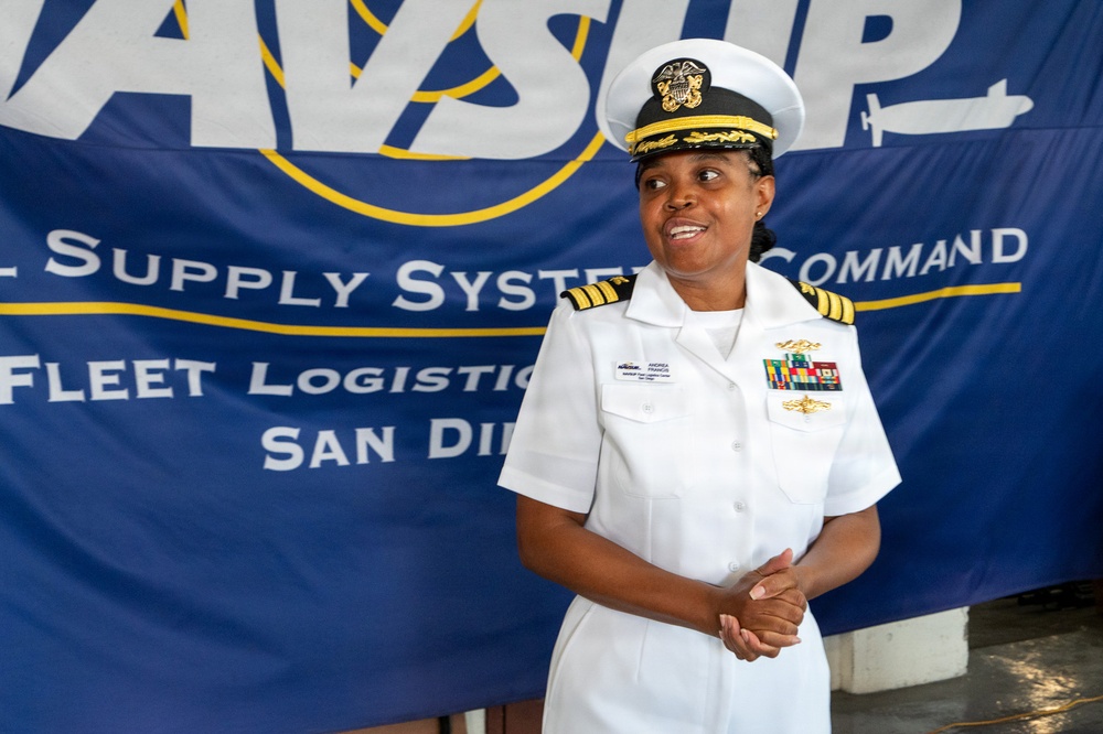 A U.S. Navy officer in white dress uniform stands in front of a Navy Supply Systems Command Fleet Logistics Center San Diego banner. The officer wears a combination cover with gold insignia, four gold sleeve stripes indicating the rank of Captain (O-6), and multiple service ribbons on the left chest. The individual appears to be speaking or presenting at an official military event.