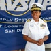 A U.S. Navy officer in white dress uniform stands in front of a Navy Supply Systems Command Fleet Logistics Center San Diego banner. The officer wears a combination cover with gold insignia, four gold sleeve stripes indicating the rank of Captain (O-6), and multiple service ribbons on the left chest. The individual appears to be speaking or presenting at an official military event.