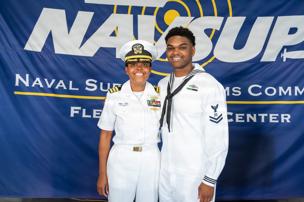 Two U.S. Navy personnel pose together in front of a NAVSUP (Naval Supply Systems Command) Fleet Logistics Center banner. The individual on the left appears to be a commissioned officer wearing a white service uniform with a combination cover, gold insignia, and multiple ribbons. The individual on the right is wearing an enlisted sailor's white dress uniform with a Dixie cup-style collar and petty officer insignia on the sleeve.