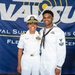 Two U.S. Navy personnel pose together in front of a NAVSUP (Naval Supply Systems Command) Fleet Logistics Center banner. The individual on the left appears to be a commissioned officer wearing a white service uniform with a combination cover, gold insignia, and multiple ribbons. The individual on the right is wearing an enlisted sailor's white dress uniform with a Dixie cup-style collar and petty officer insignia on the sleeve.