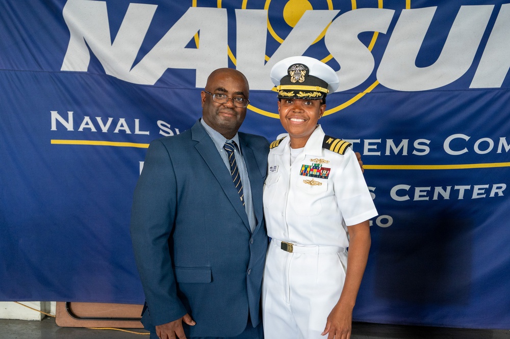 Two individuals pose for a photo in front of a Naval Supply Systems Command (NAVSUP) banner. One person is wearing a U.S. Navy officer's white dress uniform with a combination cover, rank insignia, and multiple service ribbons. The other person is dressed in a blue business suit with a striped tie.