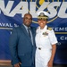 Two individuals pose for a photo in front of a Naval Supply Systems Command (NAVSUP) banner. One person is wearing a U.S. Navy officer's white dress uniform with a combination cover, rank insignia, and multiple service ribbons. The other person is dressed in a blue business suit with a striped tie.