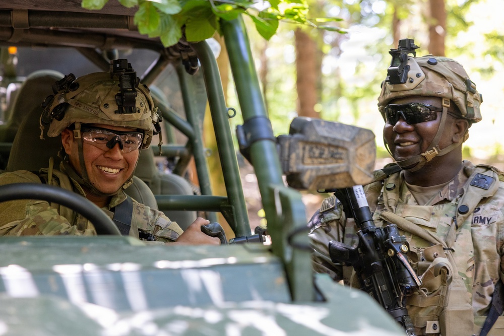 Two U.S. Army soldiers in full combat gear are interacting in a wooded outdoor setting. One soldier is seated inside a military utility vehicle (likely an M-ATV or similar light tactical vehicle), while the other stands outside holding a rifle. Both appear relaxed and are smiling, suggesting a casual moment during a training exercise or field operation.