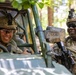 Two U.S. Army soldiers in full combat gear are interacting in a wooded outdoor setting. One soldier is seated inside a military utility vehicle (likely an M-ATV or similar light tactical vehicle), while the other stands outside holding a rifle. Both appear relaxed and are smiling, suggesting a casual moment during a training exercise or field operation.