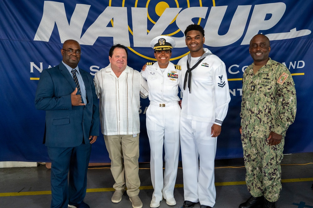 Five individuals pose for a group photo in front of a NAVSUP (Naval Supply Systems Command) banner. Three of the individuals are in U.S. Navy uniforms — one in white dress uniform with officer's cover and ribbons, one in enlisted dress whites with a Petty Officer rating badge, and one in Navy Working Uniform (NWU camouflage). Two civilians in business and casual attire complete the group.