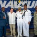 Five individuals pose for a group photo in front of a NAVSUP (Naval Supply Systems Command) banner. Three of the individuals are in U.S. Navy uniforms — one in white dress uniform with officer's cover and ribbons, one in enlisted dress whites with a Petty Officer rating badge, and one in Navy Working Uniform (NWU camouflage). Two civilians in business and casual attire complete the group.
