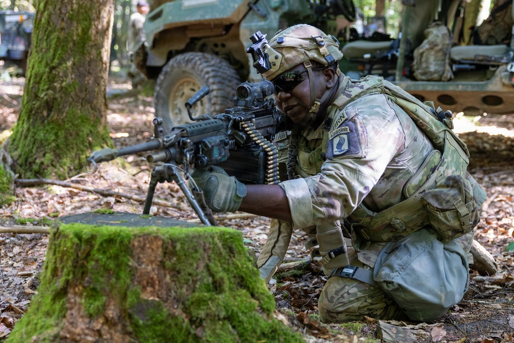 A U.S. Army Airborne soldier in full combat gear is crouched behind a moss-covered tree stump in a wooded area, aiming an M249 Squad Automatic Weapon (SAW) with a belt of ammunition loaded. The soldier wears MultiCam (OCP) camouflage, a tactical helmet with night-vision mount, and a shoulder patch identifying the 101st Airborne Division. Military vehicles are visible in the background among the trees.
