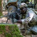 A U.S. Army Airborne soldier in full combat gear is crouched behind a moss-covered tree stump in a wooded area, aiming an M249 Squad Automatic Weapon (SAW) with a belt of ammunition loaded. The soldier wears MultiCam (OCP) camouflage, a tactical helmet with night-vision mount, and a shoulder patch identifying the 101st Airborne Division. Military vehicles are visible in the background among the trees.
