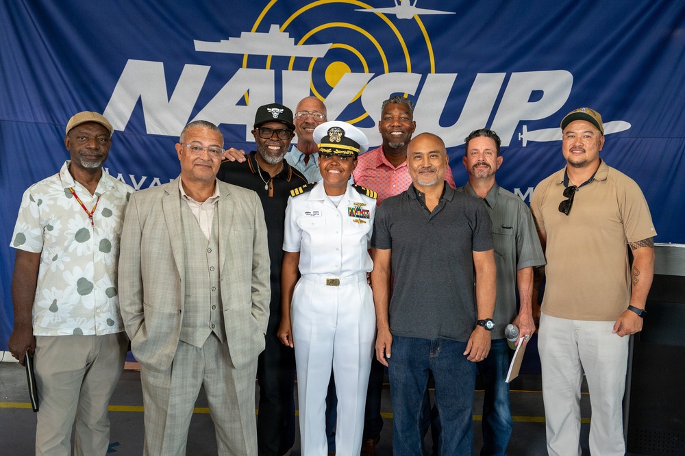 A group of approximately nine individuals pose for a formal group photo in front of a large blue 'NAVCUP' (Naval Cup) banner. One person in the center is wearing a white U.S. Navy dress uniform with officer insignia, medals, and a command cap, suggesting a military event or ceremony. The remaining individuals are dressed in civilian attire ranging from casual to business casual.